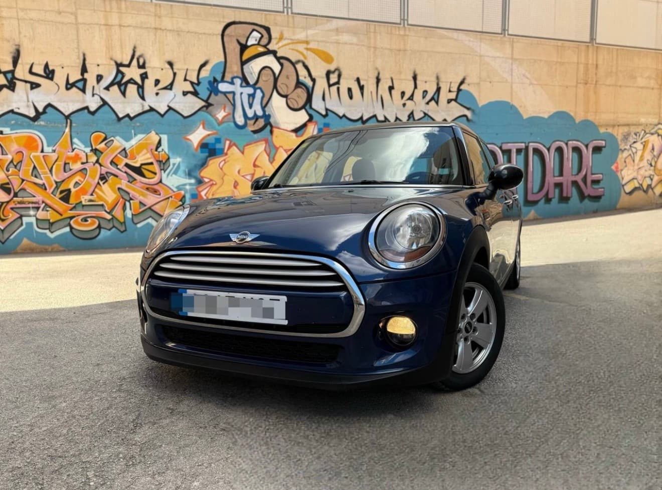Mini Cooper azul oscuro estacionado frente a una pared con grafitis de colores vibrantes.